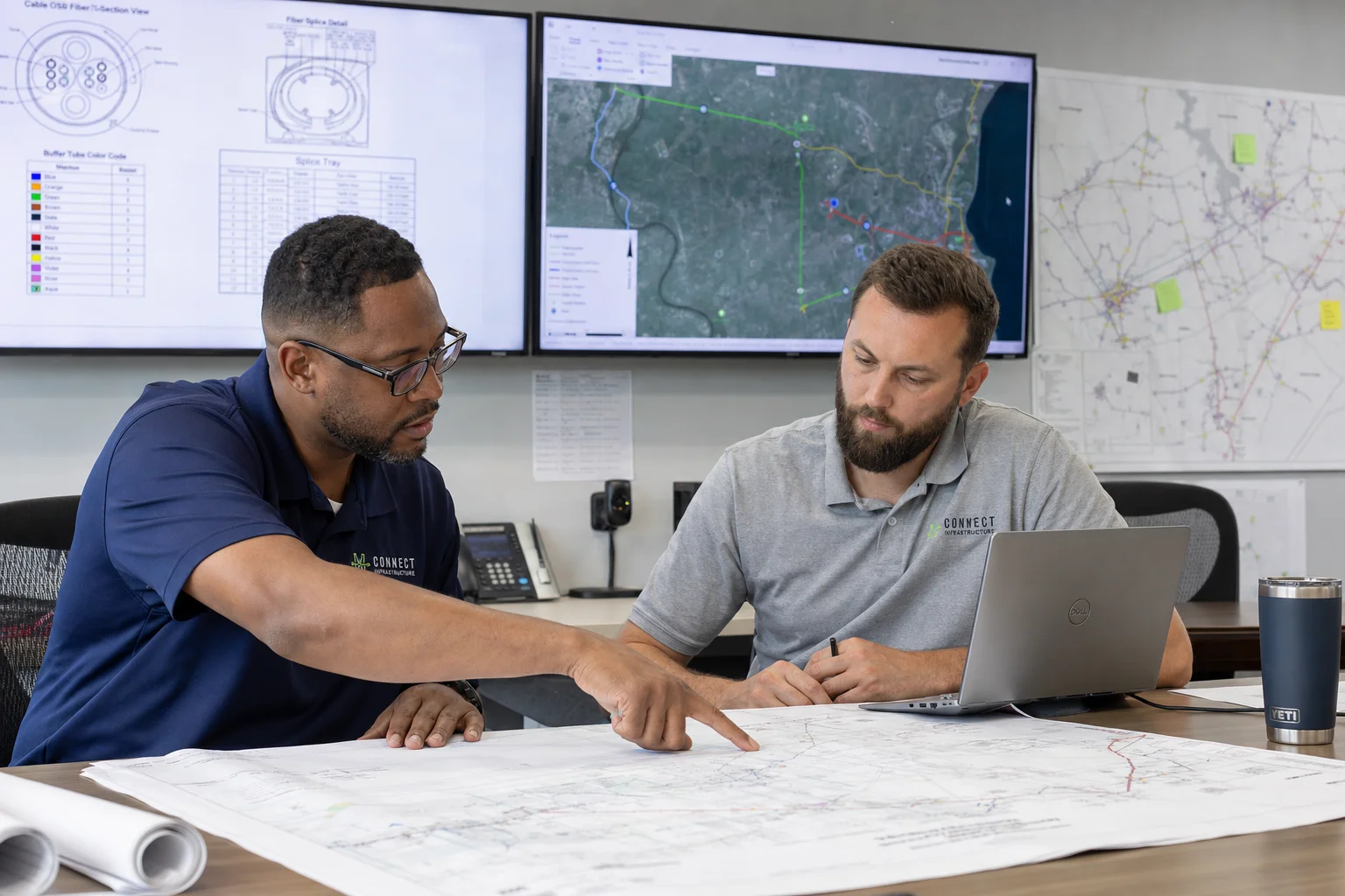OSP engineer reviewing fiber network design drawings at a workstation with GIS route maps displayed on dual monitors